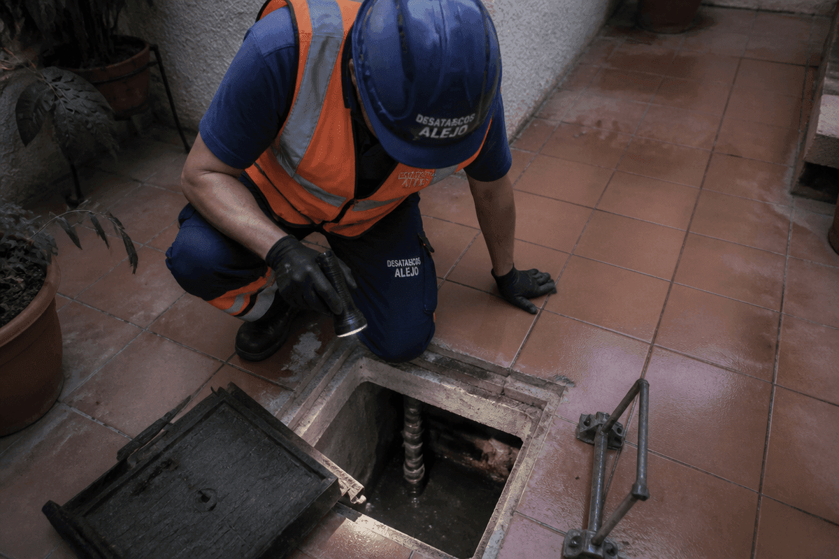 Técnico arrodillado con casco azul rotulado ilumina con linterna el interior de una arqueta cuadrada en un patio de baldosas terracota, con tapa retirada a un lado
